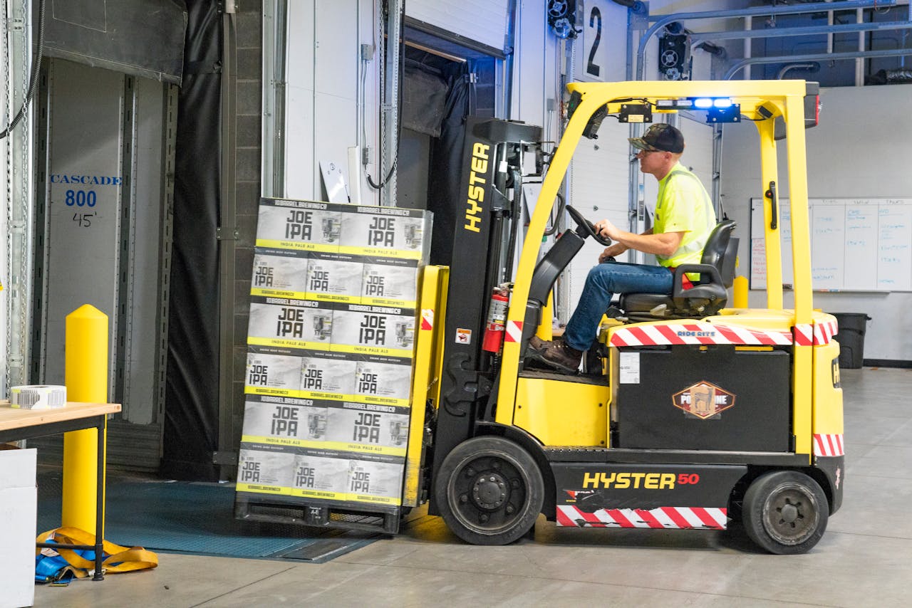 Services-03 A worker drives a Hyster forklift moving Joe IPA boxes in a warehouse.
