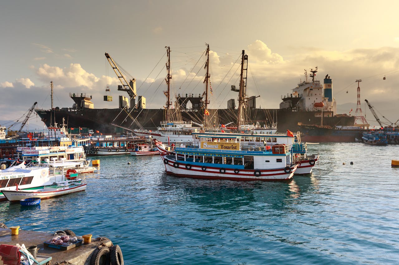 brand-01 A busy harbor with cargo ships, cranes, and tourist boats under a cloudy sky.